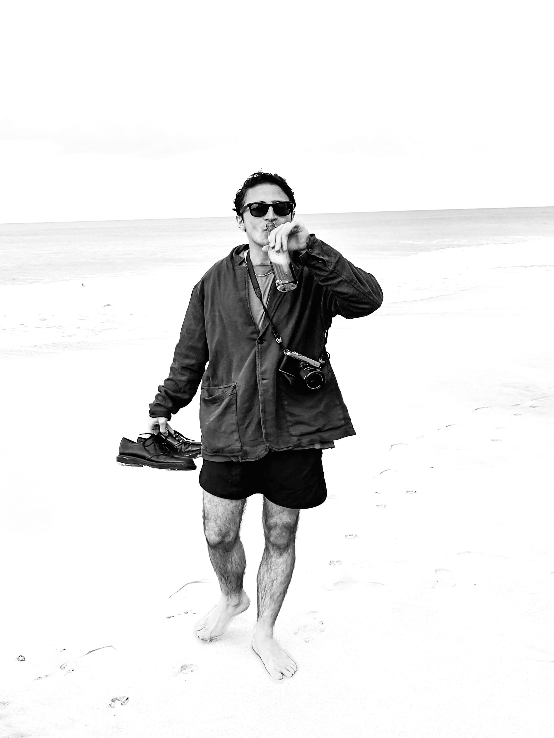 Erik Hadife walking barefoot on a beach, shoes in hand, camera around his neck.